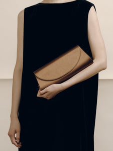 Front view of The Opera Clutch in dark brown satin showcasing its minimalist curved flap with a model in a black dress