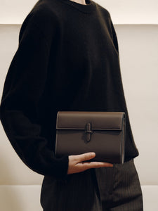 Front view of The Small Soft Clutch in Dark Brown Nappa showcasing its fold-over silhouette and slip-through closure against a black outfit