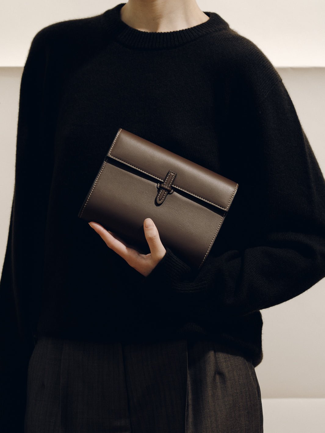 Front view of The Small Soft Clutch in dark brown Nappa leather with fold-over silhouette and slip-through closure, held by a model