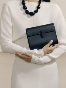 Front view of The Small Soft Clutch in navy nappa showcasing the fold-over silhouette and slip-through closure with a model in a white dress