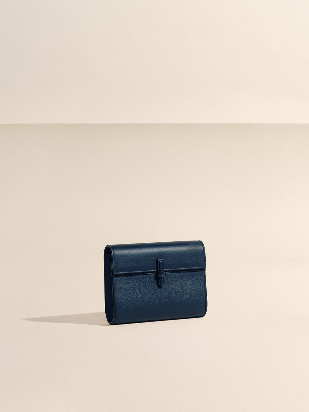 Front view of The Small Soft Clutch in Navy Nappa showcasing its fold-over silhouette and slip-through closure with a beige background