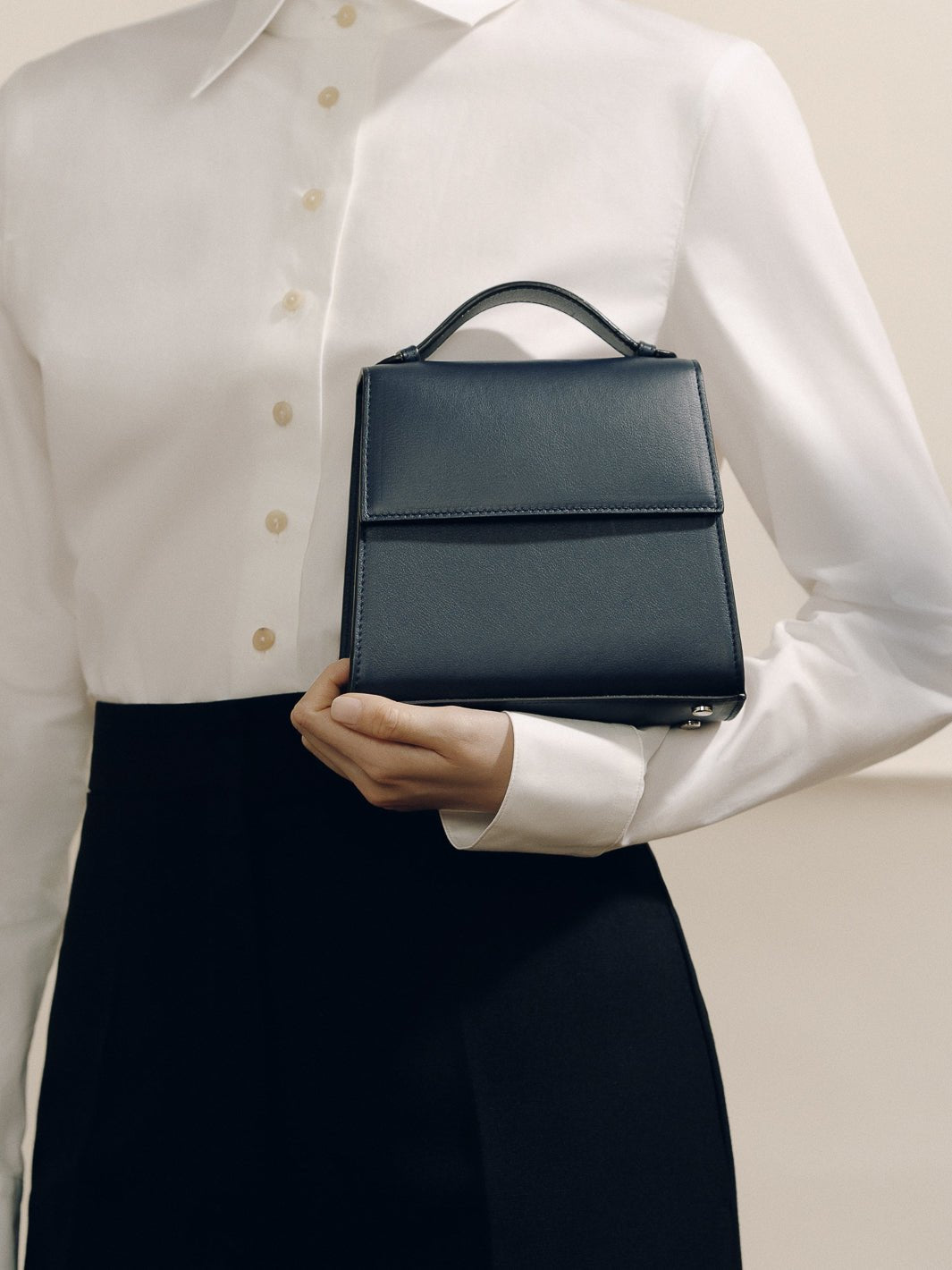 Front view of the Small Top Handle bag showcasing its rounded top handle and structured silhouette against a white blouse and black skirt