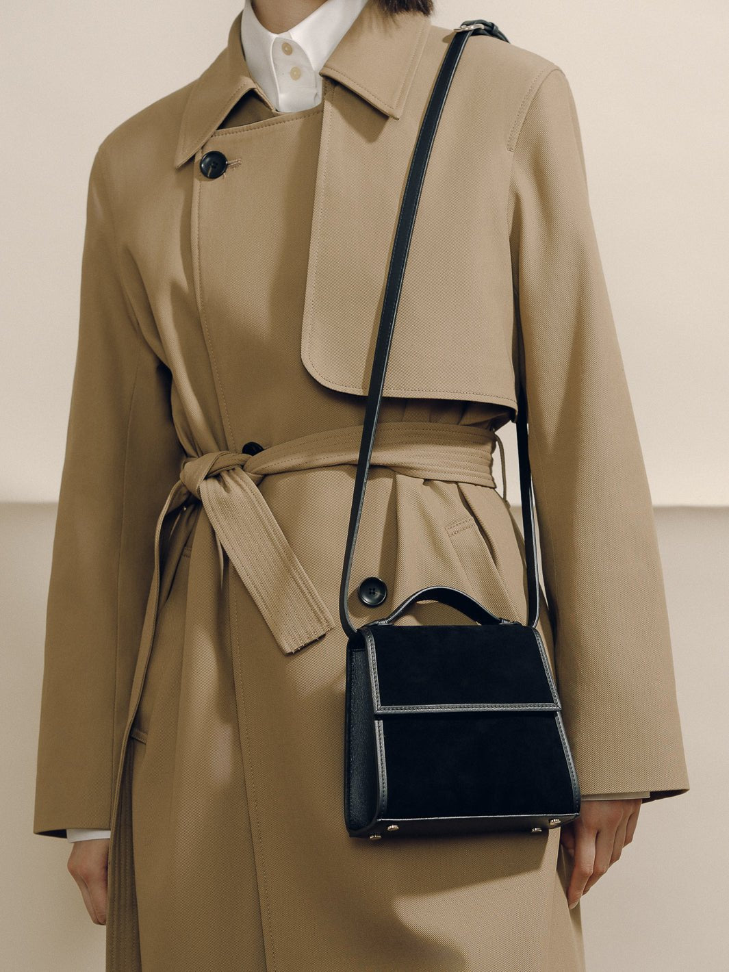 Front view of The Small Top Handle in black suede with rounded handle and adjustable strap against a beige trench coat