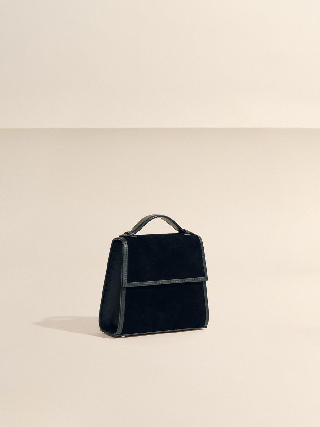 Front three quarter view of The Small Top Handle in black suede showcasing its rounded top handle against a beige background
