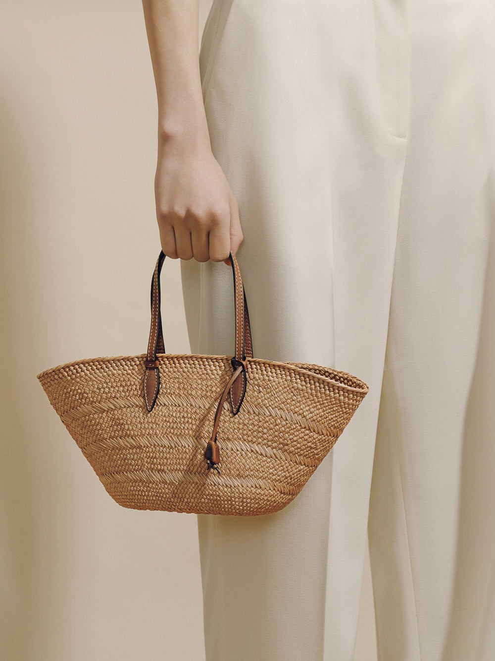 Side view of The Small Trapeze Basket in Nuez Cognac with woven body, leather handles and tagua charm against cream backdrop