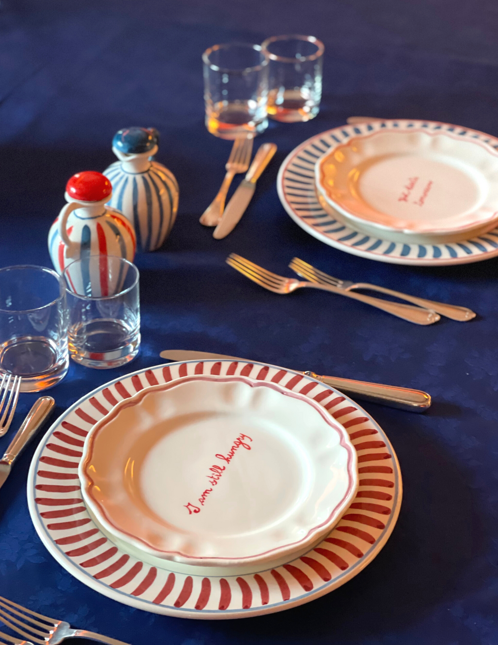 Angled view of champagne scalloped soup plate with red handwritten I am still hungry on white ceramic, on red-striped charger