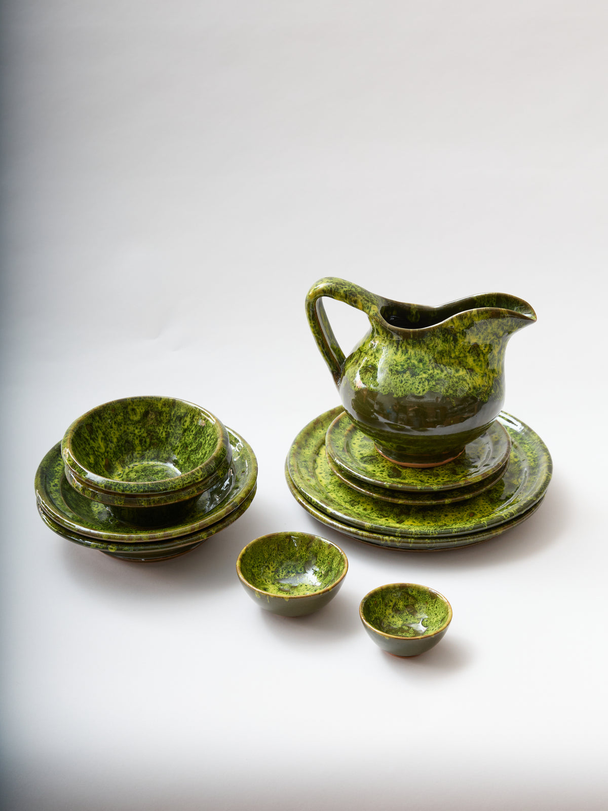 Three-quarter view of Moss terracotta pitcher showing moss-inspired green glaze with matching plates and bowls on white background
