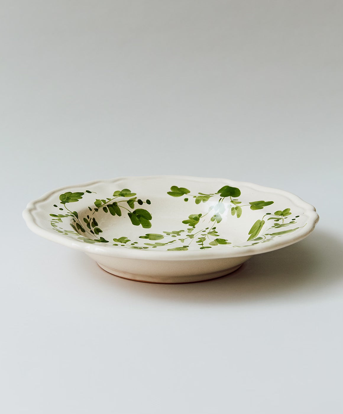 Angled view of Flora pasta bowl with hand-painted green floral motif, scalloped rim, and terracotta base
