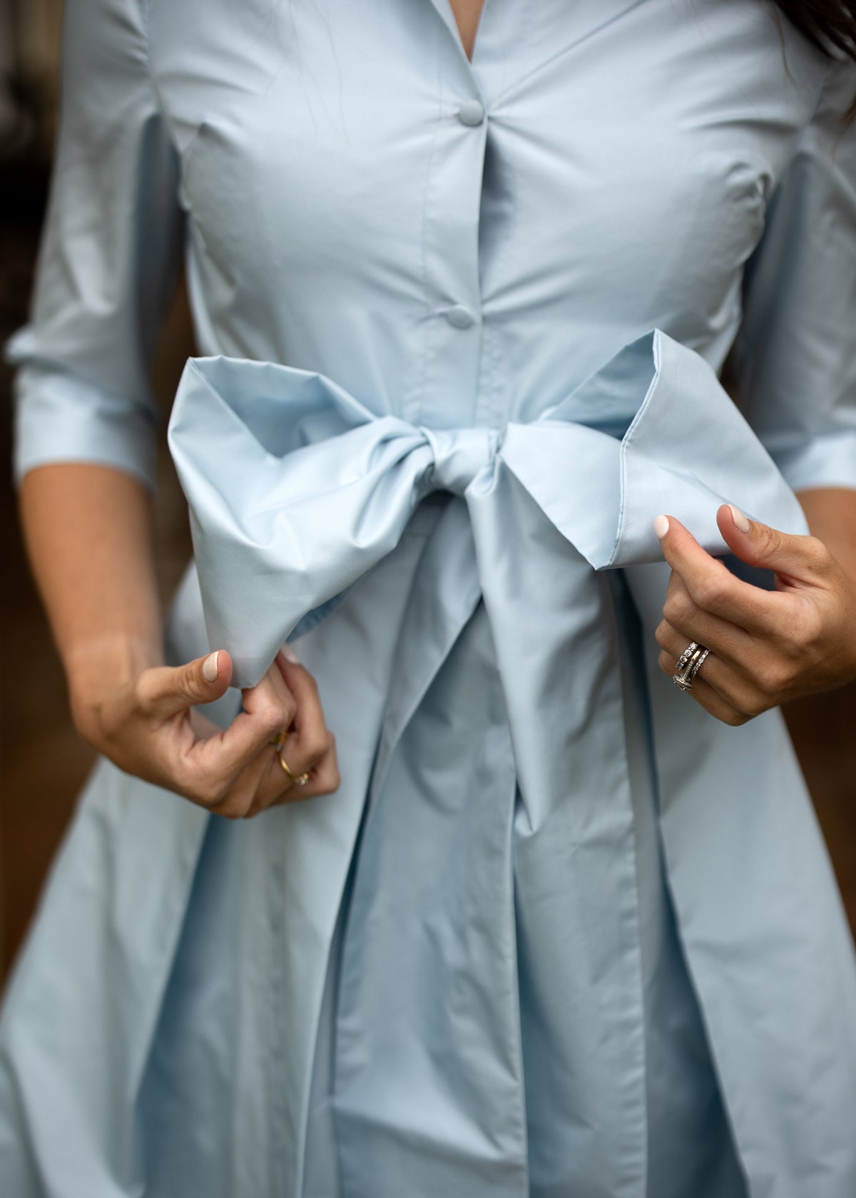 Front close-up of High Tea Dress in Blushed Blue showcasing covered buttons and bow belt with hands adjusting the dress
