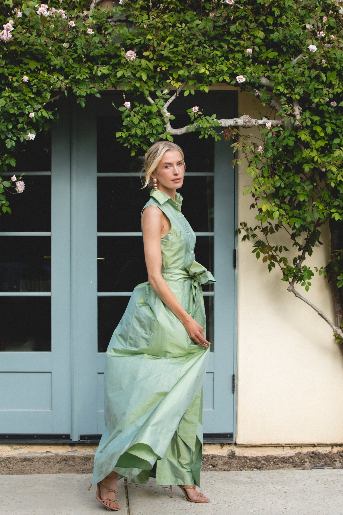 Three-quarter view of Sleeveless Collar Gown in Soft Sage showcasing its bow belt and train by a rose-covered doorway