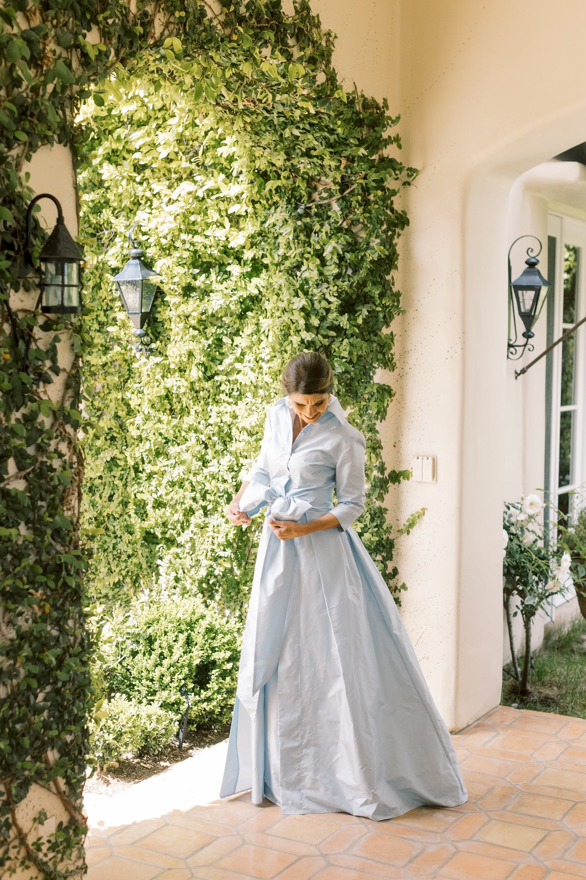 Front view of Effortlessly Elegant Gown in Blushed Blue showcasing collared neckline and bow belt in a leafy garden