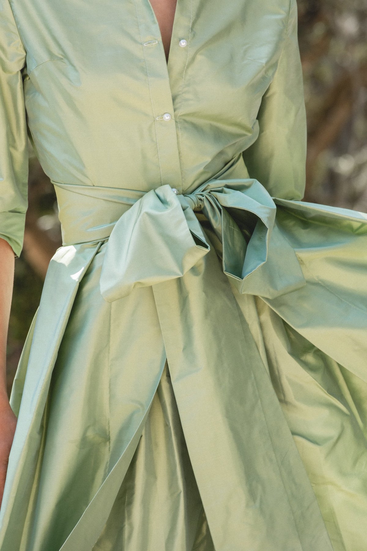 Close-up of Effortlessly Elegant Gown in soft sage showcasing tied waist bow, pearl-button front, and voluminous skirt outdoors