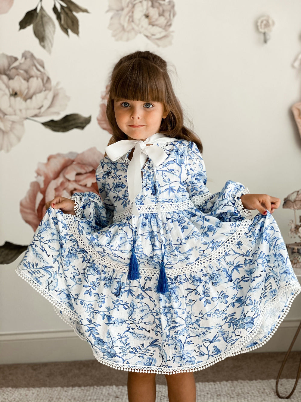 Front view of Jaylin linen dress showcasing blue and white floral print, puff sleeves, lace trim and tassel ties against floral wallpaper