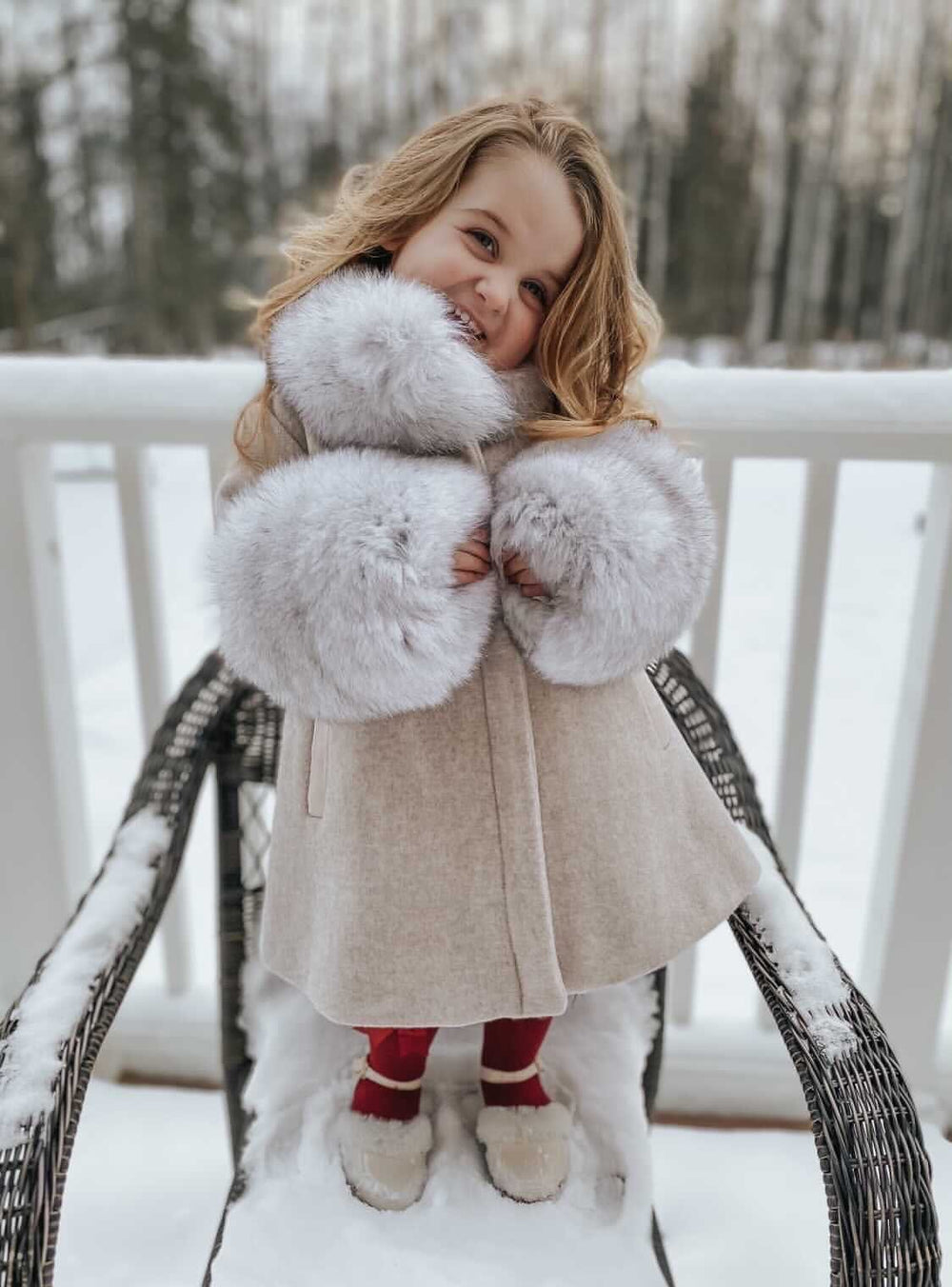 Front view of Karenina wool coat in light beige on a child model, showing oversized grey fur collar and cuffs on a snowy porch