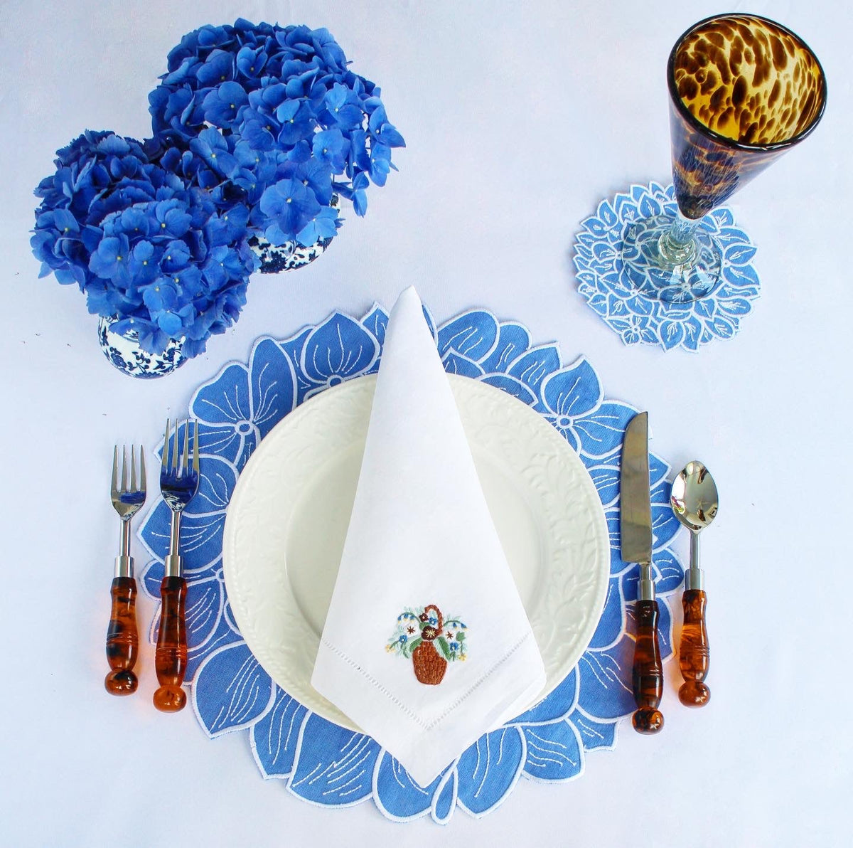 Overhead view of the Hydrangea Placemat showcasing blue linen, white-thread floral details, and a set table background