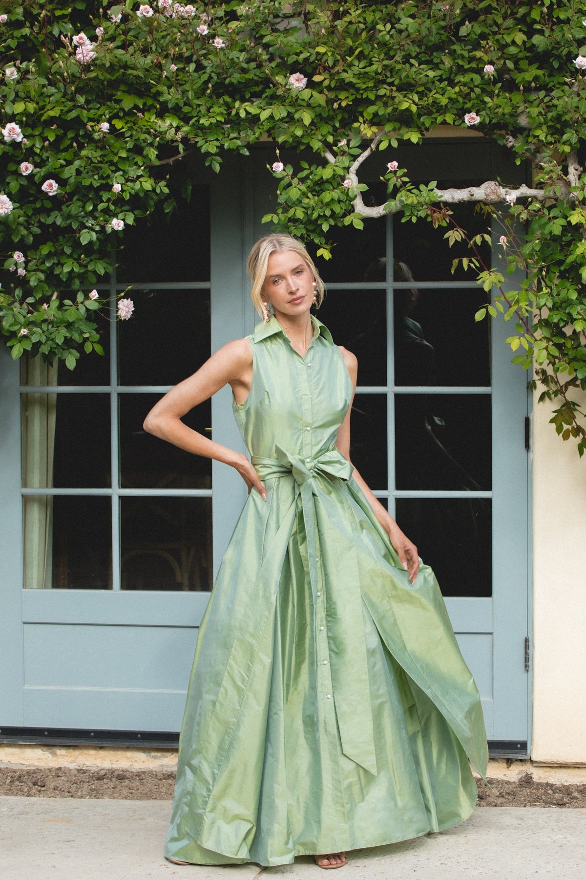 Front view of Sleeveless Collar Gown in Soft Sage showcasing collar, bow belt, and full skirt against a garden window
