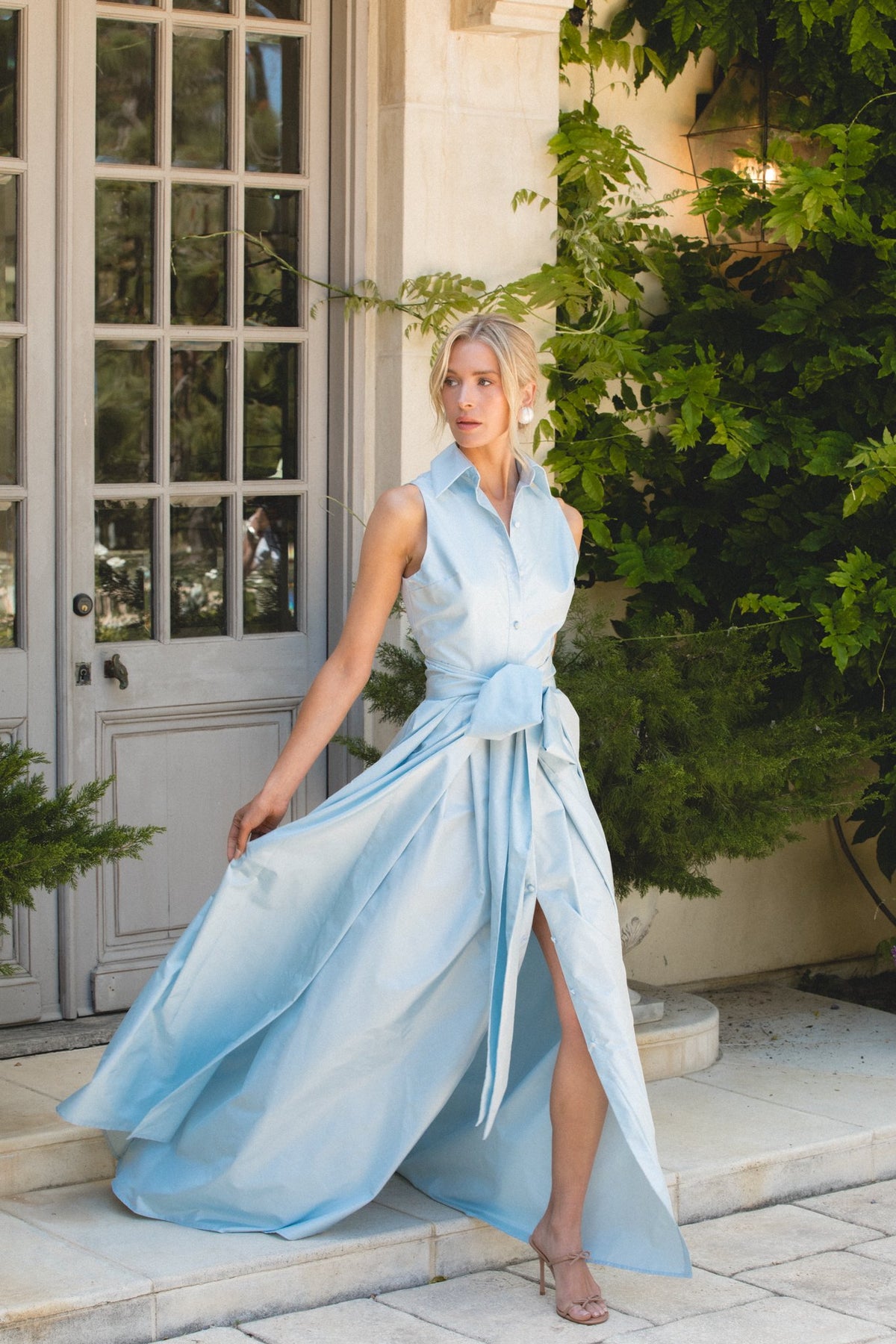Front view of sleeveless collared gown in blushed blue showcasing tied waist bow, high side slit, and flowing skirt