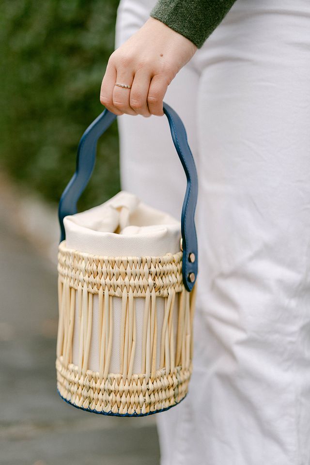 Handheld Emily Tote showing woven reed body, navy leather handle and canvas insert, blurred background