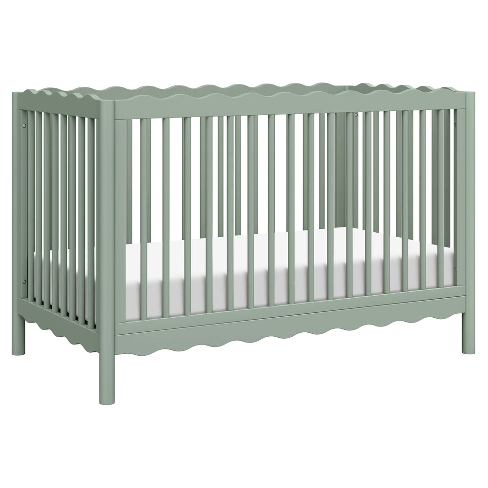 Three-quarter view of Swell 4-in-1 convertible crib showing scalloped rails, vertical slats, and light sage finish on white