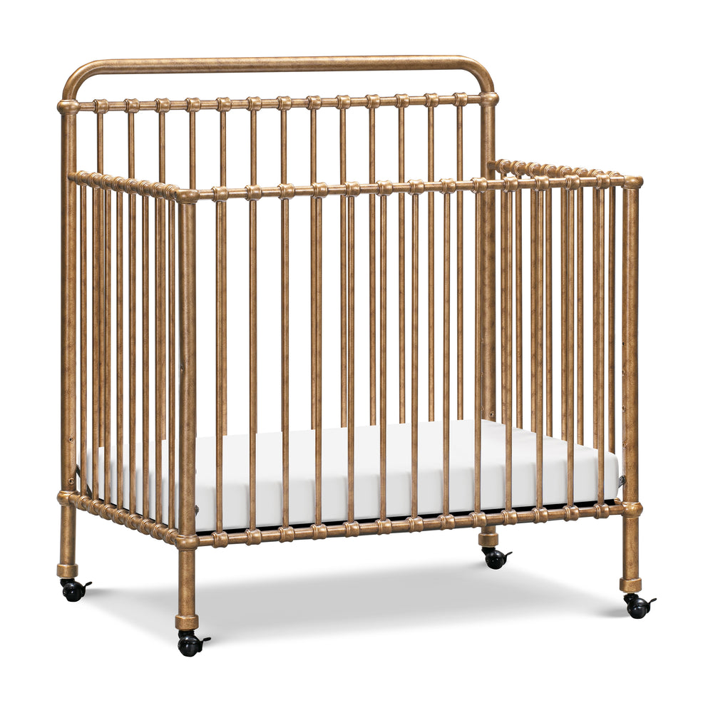 Angled view of Winston 4-in-1 mini crib in vintage gold showing metal spindle frame and caster wheels