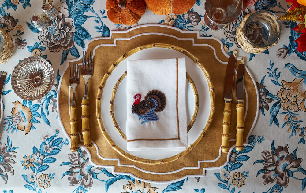 Top-down view of Grace placemat showcasing scalloped mustard linen with white embroidered border on floral tablecloth