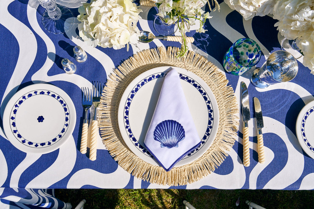 Overhead view of Siena Coquillage Napkins, Set Of 4 showcasing a clam shell and navy trim on a blue and white table setting