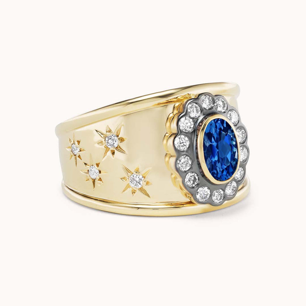 Angled view of Ali Cigar Band in 14K yellow gold showing oval blue sapphire with diamond halo and starburst diamond accents