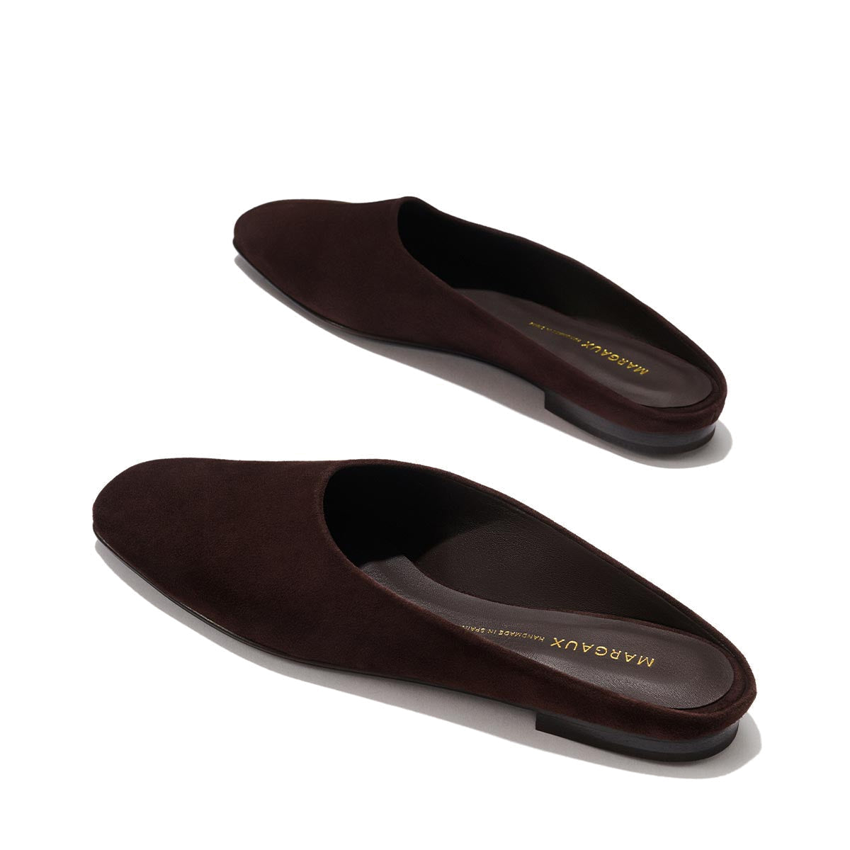 The Clara - Espresso Suede by Margaux are dark brown suede slip-on mules with flat soles and a minimalist, slipper-inspired design. Shown on a light background, the brand name Margaux is visible inside the shoes.