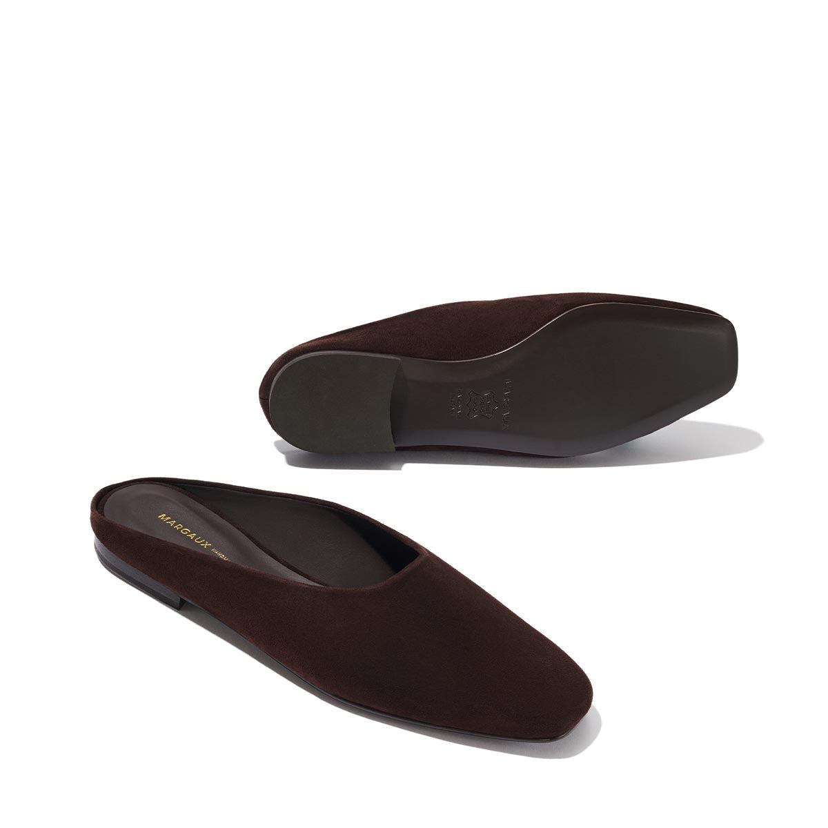 The Clara - Espresso Suede by Margaux: dark brown suede slip-ons with a slipper-like silhouette, squared toes, flat soles, and a minimalist design. One shoe is upright; the other lies on its side to show the sole.