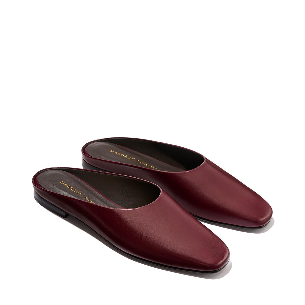 A pair of The Clara - Wine Calf slip-on mules by Margaux, crafted in rich burgundy leather with a subtle heel and displayed side by side on a neutral background, these shoes effortlessly blend slipper-like comfort with elevated chic for versatile, do-it-all style.