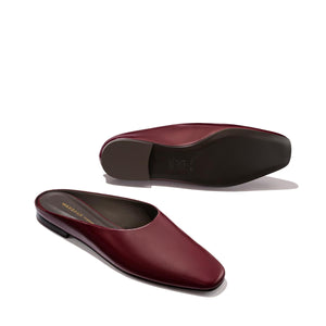 A pair of Margaux's The Clara - Wine Calf leather slip-on flats with a slipper silhouette, closed toes, and open backs, displayed on a light background. One shoe stands upright while the other lies on its side to showcase the sole.