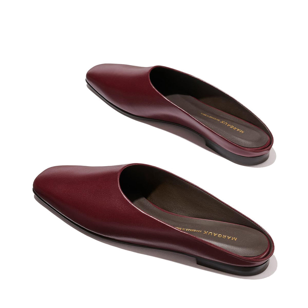 Introducing The Clara - Wine Calf by Margaux: a pair of versatile burgundy leather slip-on mules featuring square toes and low black heels in a classic slipper silhouette, elegantly displayed on a light background.
