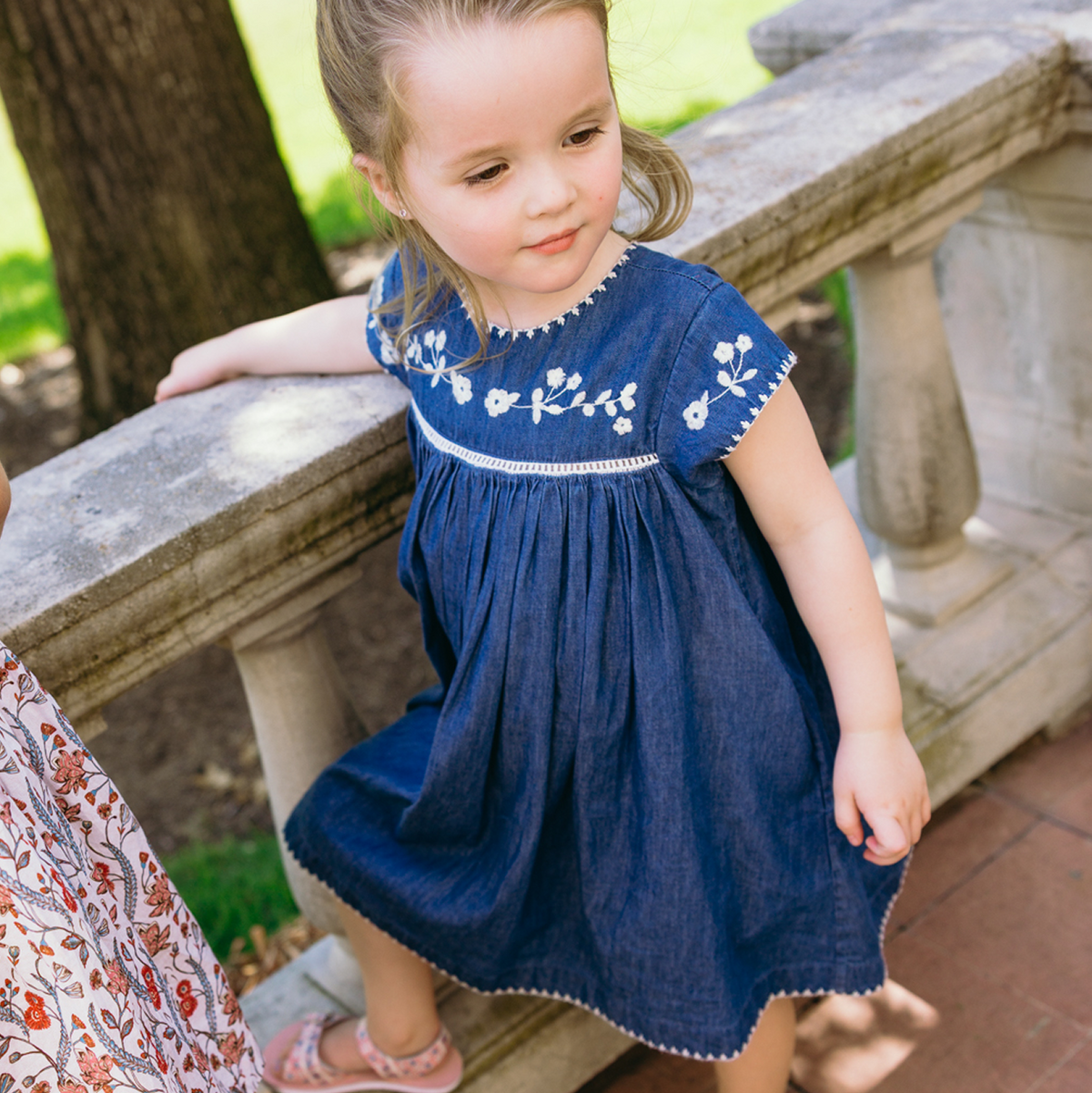 Three-quarter view of girls’ chambray dress with white floral embroidery, cap sleeves, gathered skirt, and scalloped hem by a stone railing