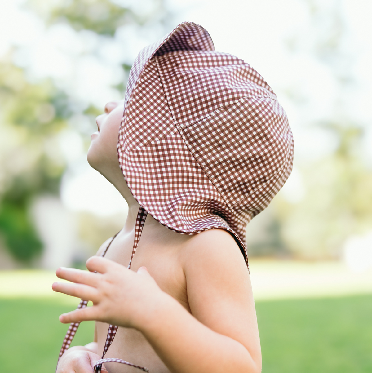 Side view of Baby Cocoa Gingham Sun Hat on a baby, showing wide brim and thick ties with blurred grassy background
