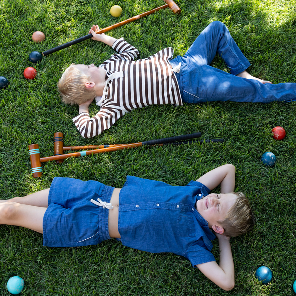 Boy wearing chambray short-sleeve button-down shirt lying on grass beside croquet mallets and balls