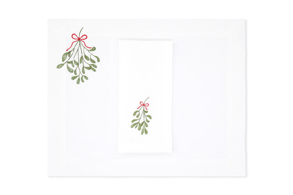 Top view of Mistletoe Collection placemat and napkin set showing green embroidered mistletoe with red bows on white linen