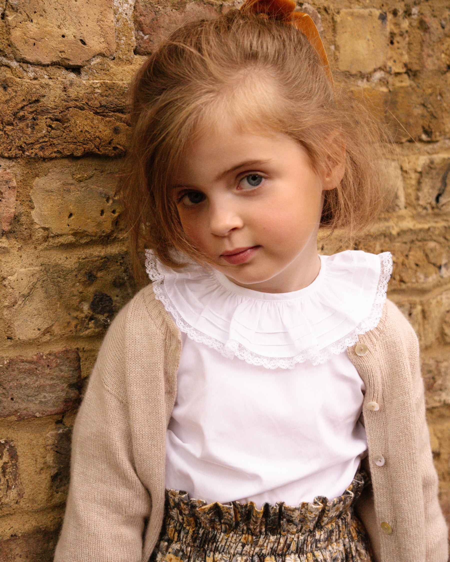 Young girl standing against a brick wall wearing a white blouse with ruffles and a beige cardigan.