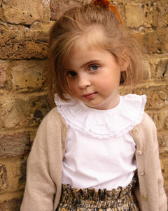 Young girl standing against a brick wall wearing a white blouse with ruffles and a beige cardigan.