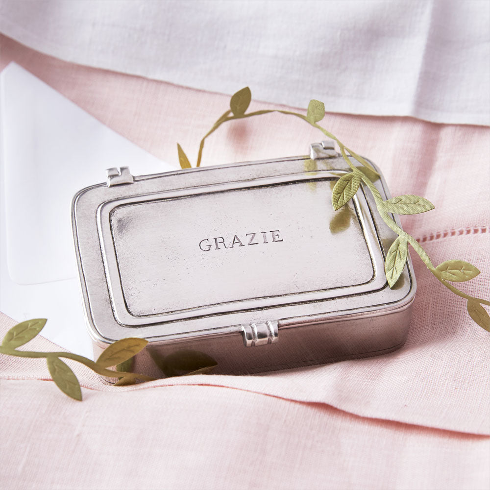Angled view of Small Grazie pewter box showcasing engraved GRAZIE lid on folded pink linen background