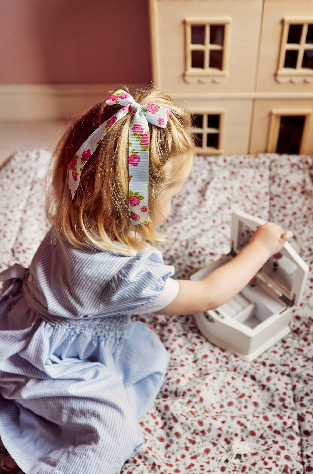 Little girl opening a blue floral musical ballerina jewelry box on a floral blanket with a wooden dollhouse in the background