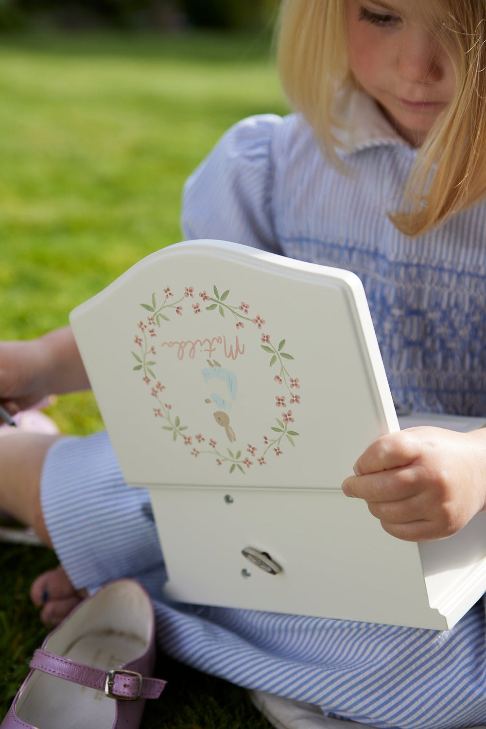 Young girl opening a hand-painted musical ballerina jewelry box with Cosima Rabbit illustration while sitting on grass