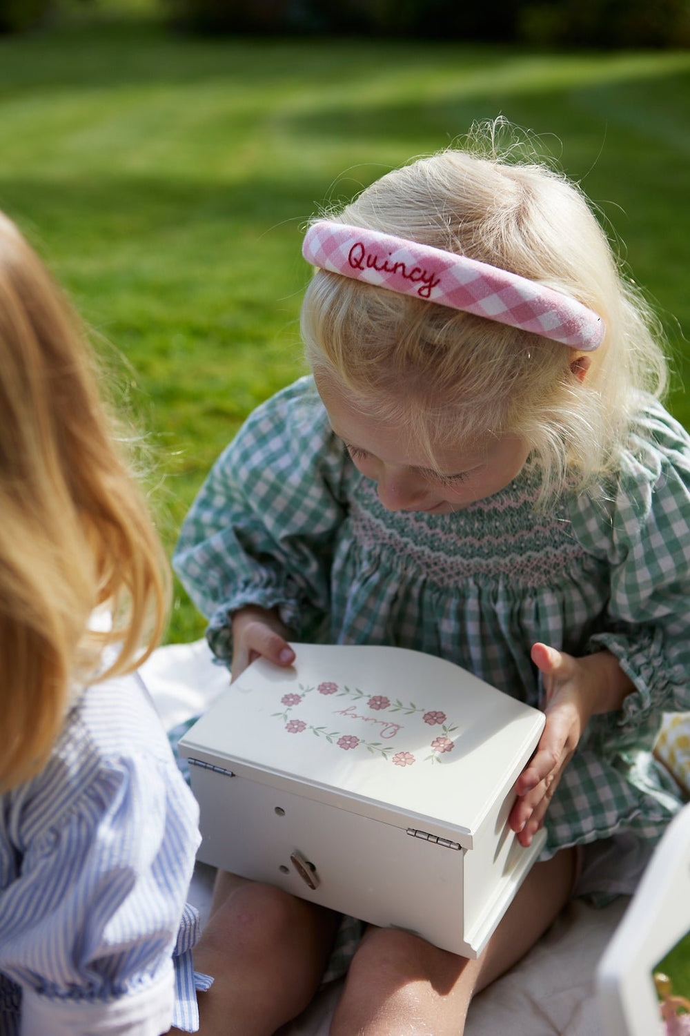 Young girl opening a white musical ballerina jewelry box with pink floral lid while sitting on a blanket outdoors