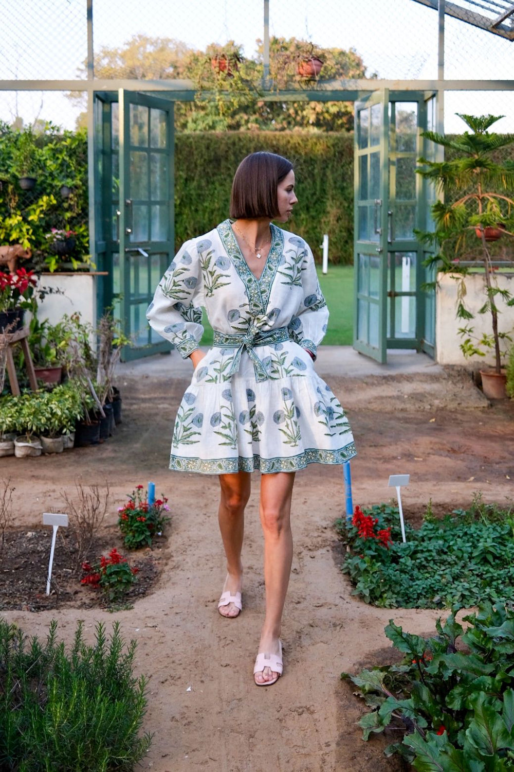 Three-quarter view of Mykonos mini dress in blue flower with v-neck, tie waist, puff sleeves, and tiered skirt in greenhouse