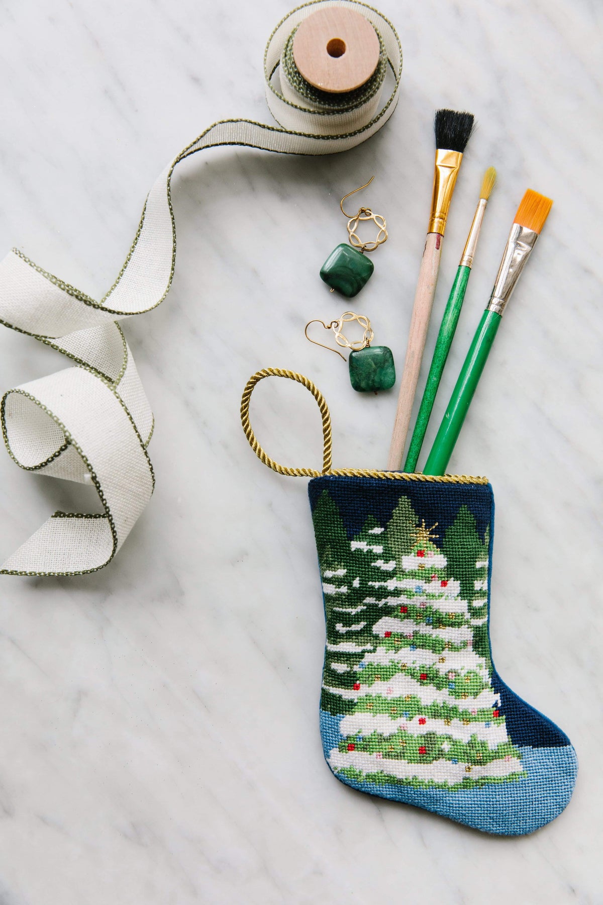 Top view of needlepoint Winter Wonderland bauble stocking with embroidered tree, gold hanging loop, and paintbrushes on marble