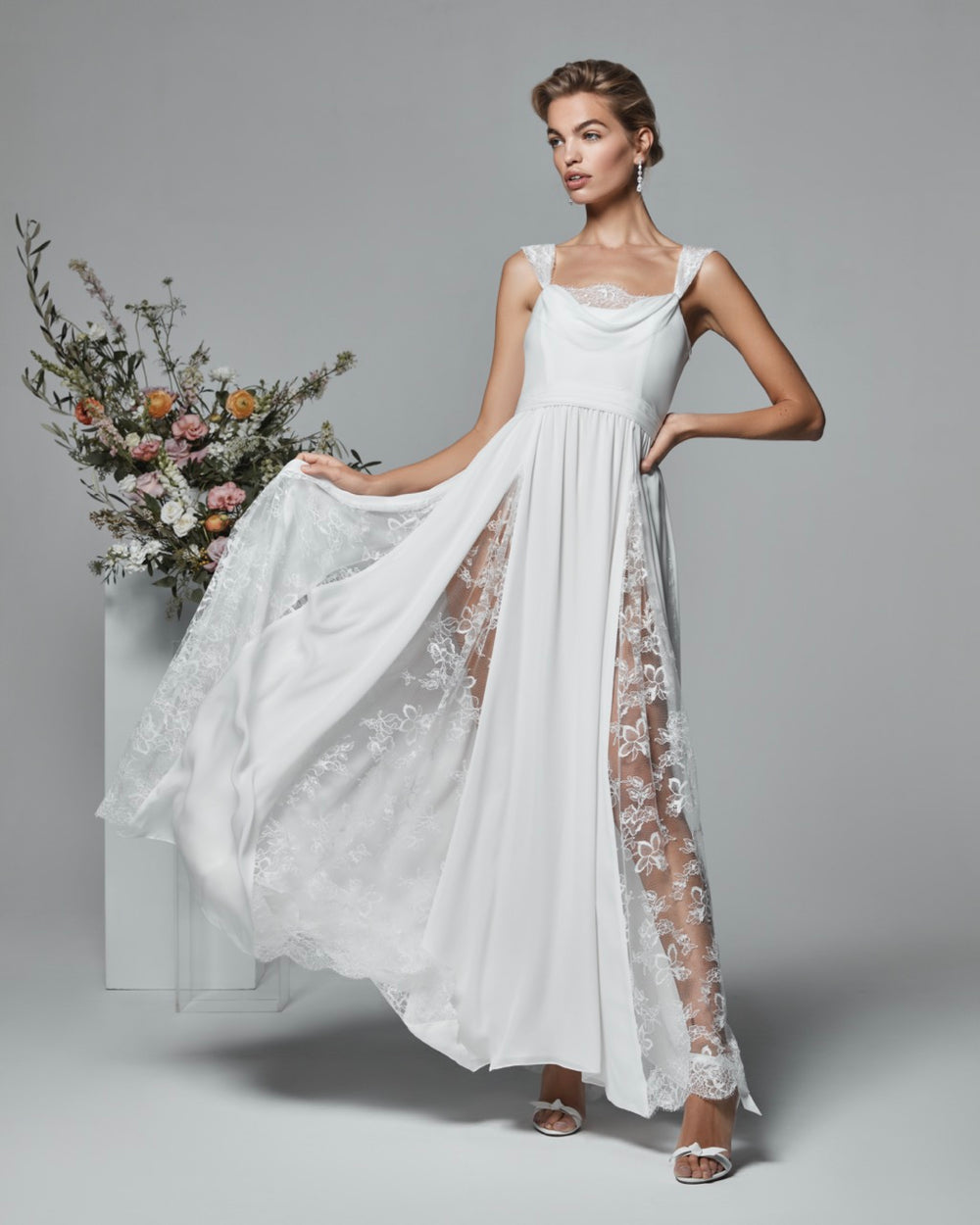 Three-quarter view of The Ivana Gown on a model showcasing cowl neckline, lace godet panels and side slit on gray background