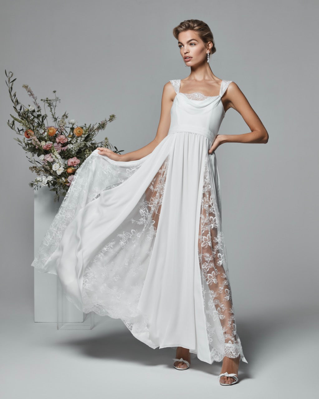 Three-quarter view of The Ivana Gown on a model showcasing cowl neckline, lace godet panels and side slit on gray background