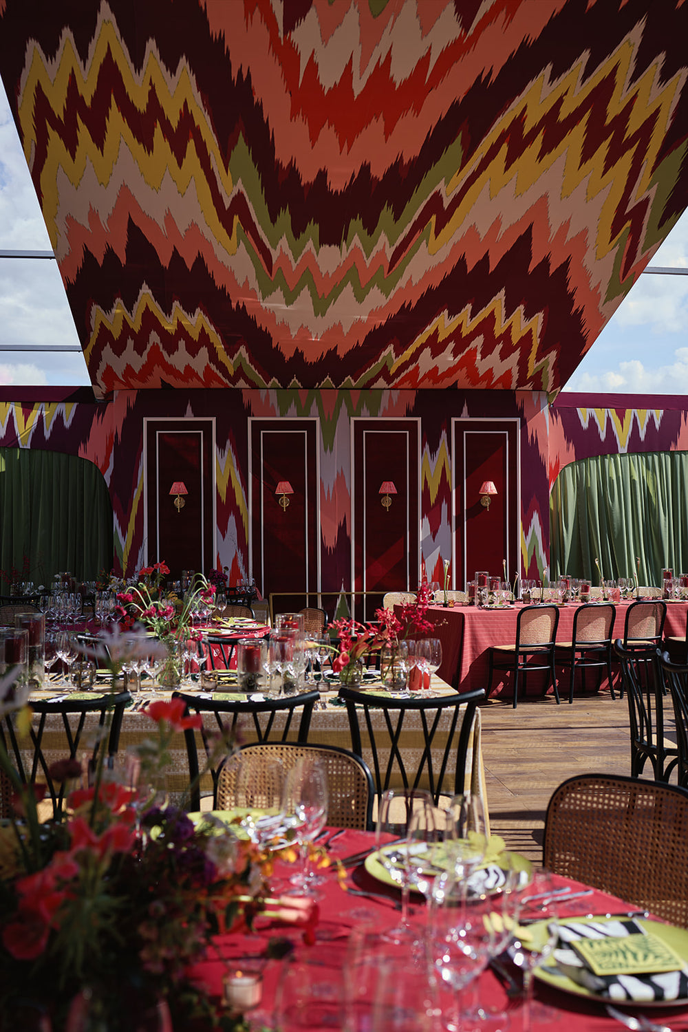 Wedding reception dining area under a colorful patterned canopy, with red tables, flowers, and place settings