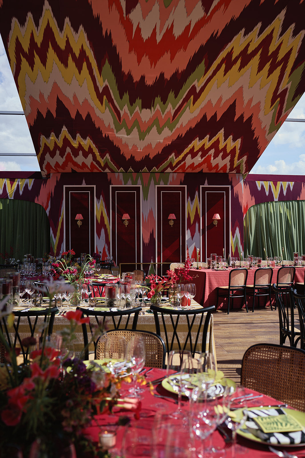 Wedding reception dining area under a colorful patterned canopy, with red tables, flowers, and place settings