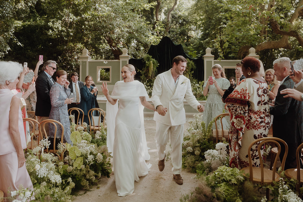 Bride and groom walk through a flower-lined garden at their Sintra wedding as guests applaud and take photos