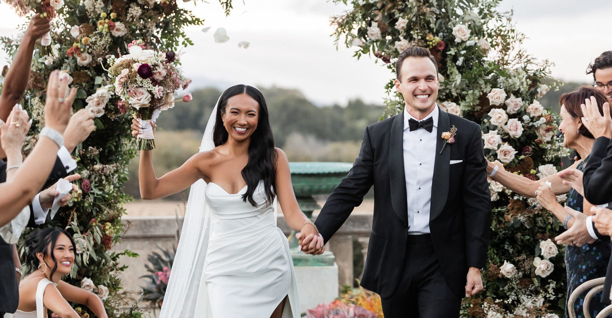 Bride and groom walk hand in hand beneath a floral wedding arch as guests celebrate outdoors